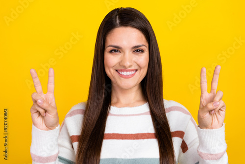 Portrait photo of brunette hair charming glad middle age lady wearing striped pullover smiling and showing v sign isolated on yellow color background