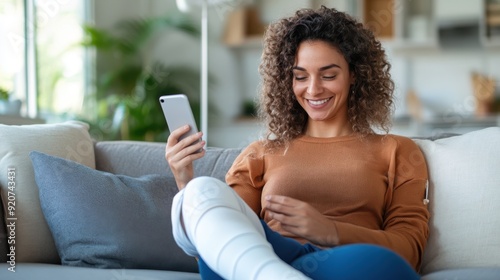 A smiling woman with curly hair relaxes on a couch holding her phone, her leg thoughtfully wrapped in a cast, indicating a scene of recovery and digital engagement indoors. photo