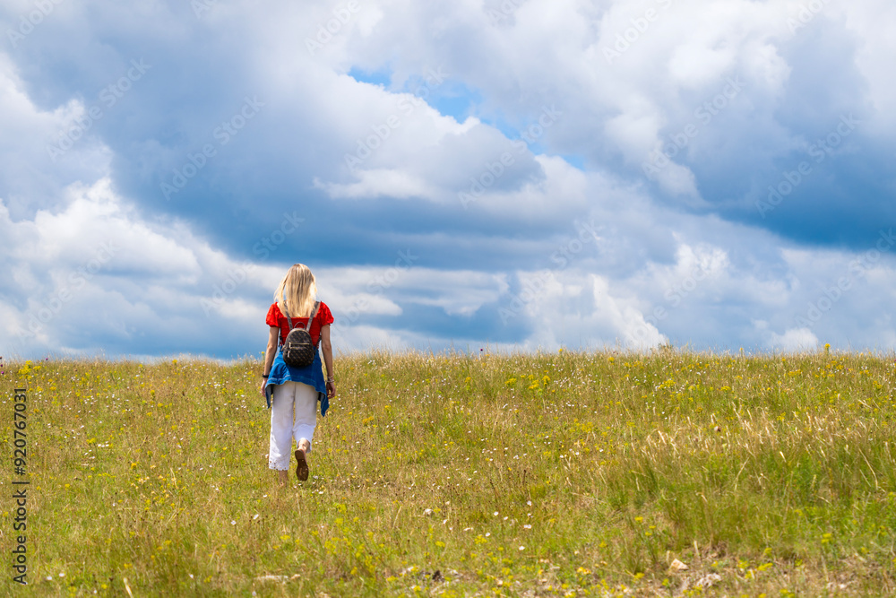 Blonde woman in red and white dress walking on a green meadow towards the cloudy horizon Blonde woman in red and white dress walking on a green meadow towards the cloudy horizon