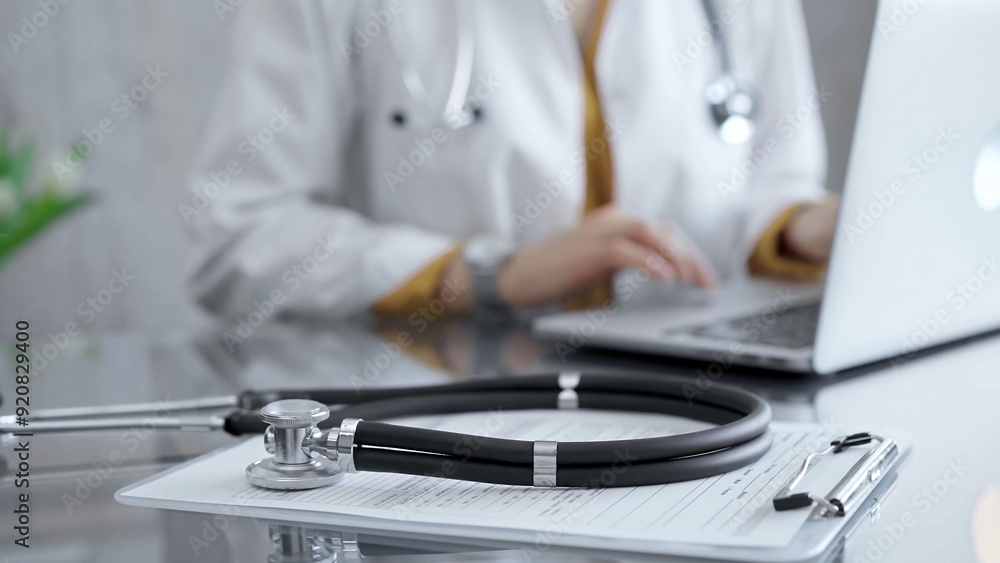 Stethoscope and clipboard with medication history records are on a glass desk behind a physician using a laptop computer. Medicine concept Stethoscope and clipboard with medication history records are on a glass desk behind a physician using a laptop computer. Medicine concept