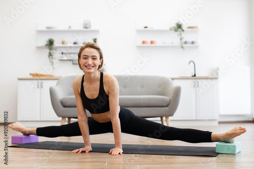 Smiling woman performing yoga split in living room on yoga mat using yoga blocks. Practicing flexibility and fitness at home with focus on stretching and balance.