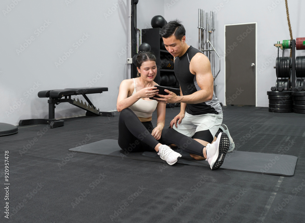 Obraz premium Young asian female sitting on the rubber floor in the gym, check the exercise schedule through tablet computer with male instructor.