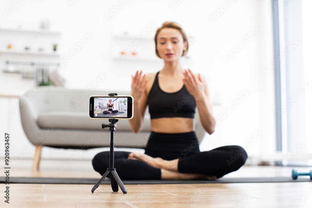 Woman in black sportswear recording online yoga class using smartphone on tripod from home. Indoor fitness training, mindfulness and healthy lifestyle concepts depicted in modern living room setting. Woman in black sportswear recording online yoga class using smartphone on tripod from home. Indoor fitness training, mindfulness and healthy lifestyle concepts depicted in modern living room setting.