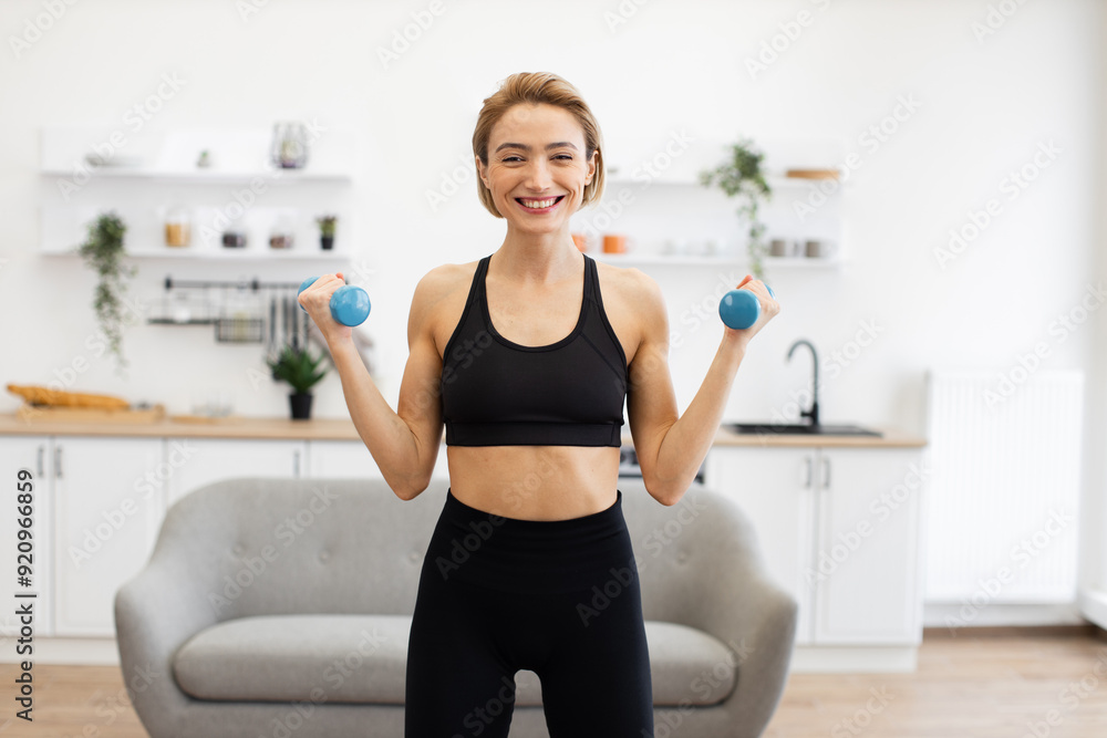 Smiling woman performing fitness exercise with blue dumbbells in contemporary kitchen. Home workout, healthy lifestyle, and strength training concept. Smiling woman performing fitness exercise with blue dumbbells in contemporary kitchen. Home workout, healthy lifestyle, and strength training concept.