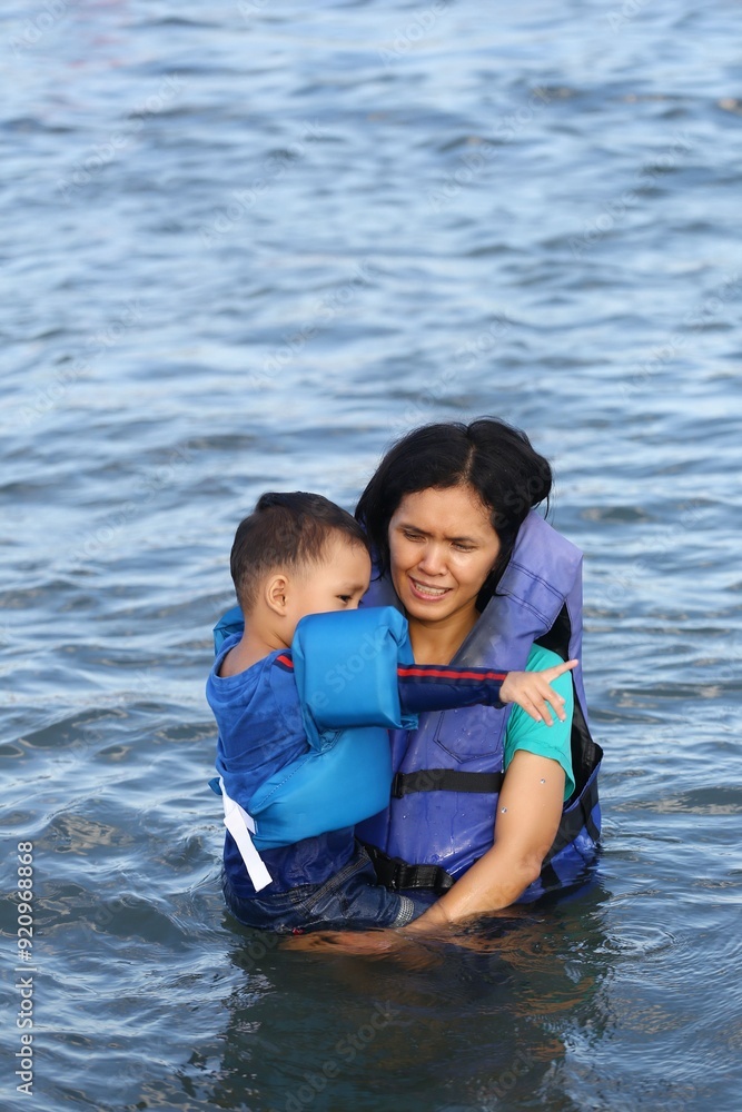 Obraz premium A asian mother holds her son, they are ware a life jacket while playing in the sea.