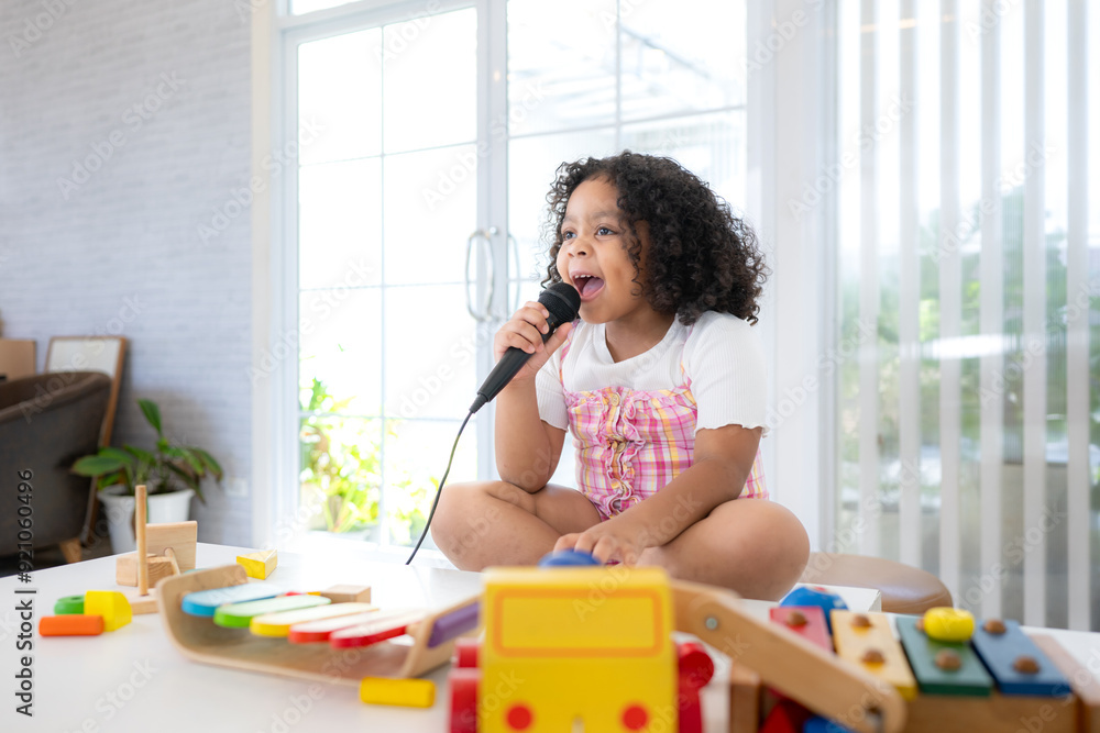 A tiny girl sits on a toy table, clutching a microphone, and sings cheerfully in the living room A tiny girl sits on a toy table, clutching a microphone, and sings cheerfully in the living room