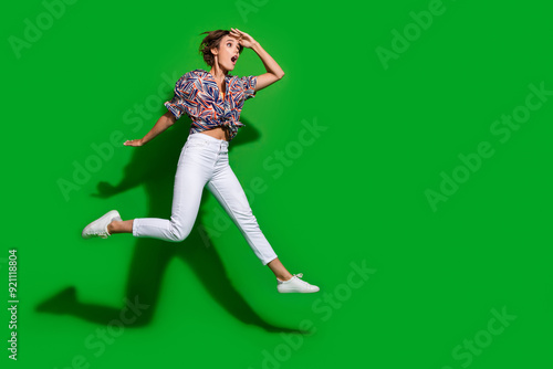 Full size photo of impressed girl wear print shirt jumping hold arm on forehead look empty space isolated on green color background