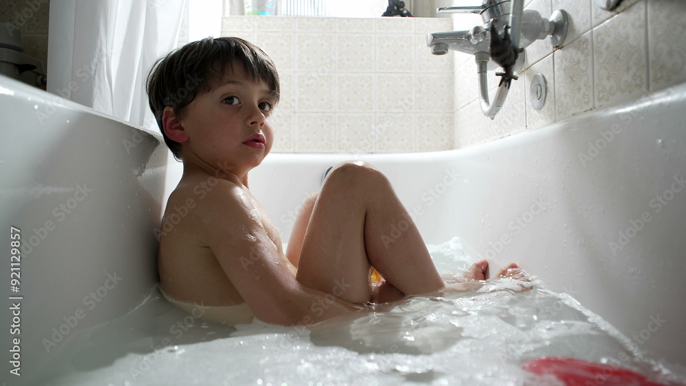 Young boy sitting in the bathtub, surrounded by water and bubbles, looking curiously at something outside the frame. candid moment of bath time Young boy sitting in the bathtub, surrounded by water and bubbles, looking curiously at something outside the frame. candid moment of bath time