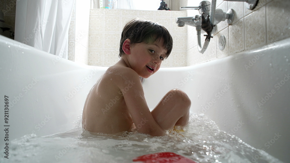 Young boy sitting in the bathtub, surrounded by water and bubbles, looking curiously at something outside the frame. candid moment of bath time Young boy sitting in the bathtub, surrounded by water and bubbles, looking curiously at something outside the frame. candid moment of bath time