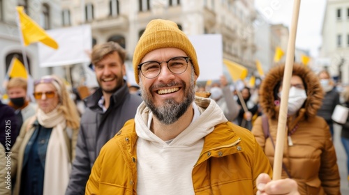 A bearded man in a yellow beanie and glasses smiles cheerfully at an outdoor protest amid a diverse and engaged crowd holding signs and banners. photo