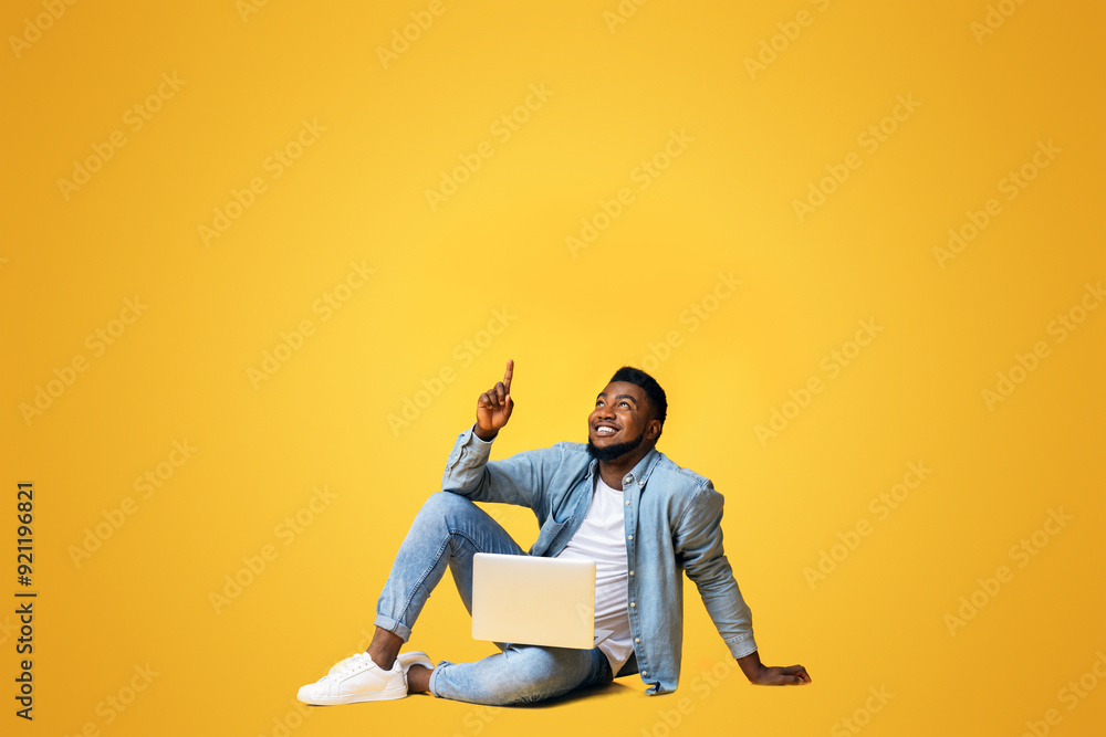 Great offer. Black millennial guy sitting on floor with laptop and pointing upwards at copy space on yellow background Great offer. Black millennial guy sitting on floor with laptop and pointing upwards at copy space on yellow background