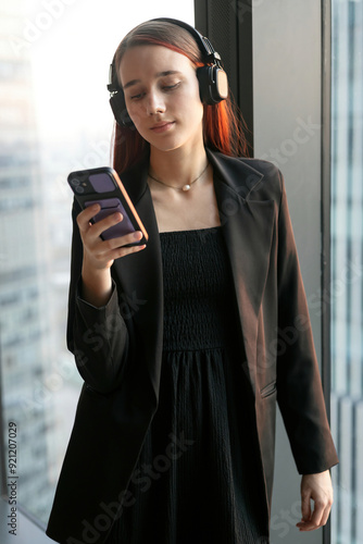 Canvastavla Young woman wearing black business suit with headphones stands at the large window of skyscraper, looking at smartphone and smiling