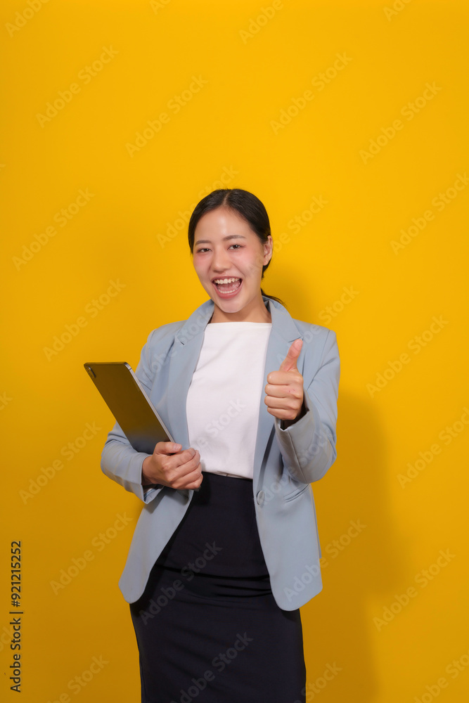Young asian businesswoman holding a tablet is smiling and showing thumbs up while standing on a yellow background Young asian businesswoman holding a tablet is smiling and showing thumbs up while standing on a yellow background