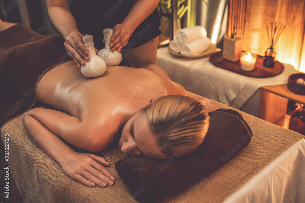Obraz premium Hot herbal ball spa massage body treatment, masseur gently compresses herb bag on woman body. Tranquil and serenity of aromatherapy recreation in warm lighting of candles at spa salon. Quiescent