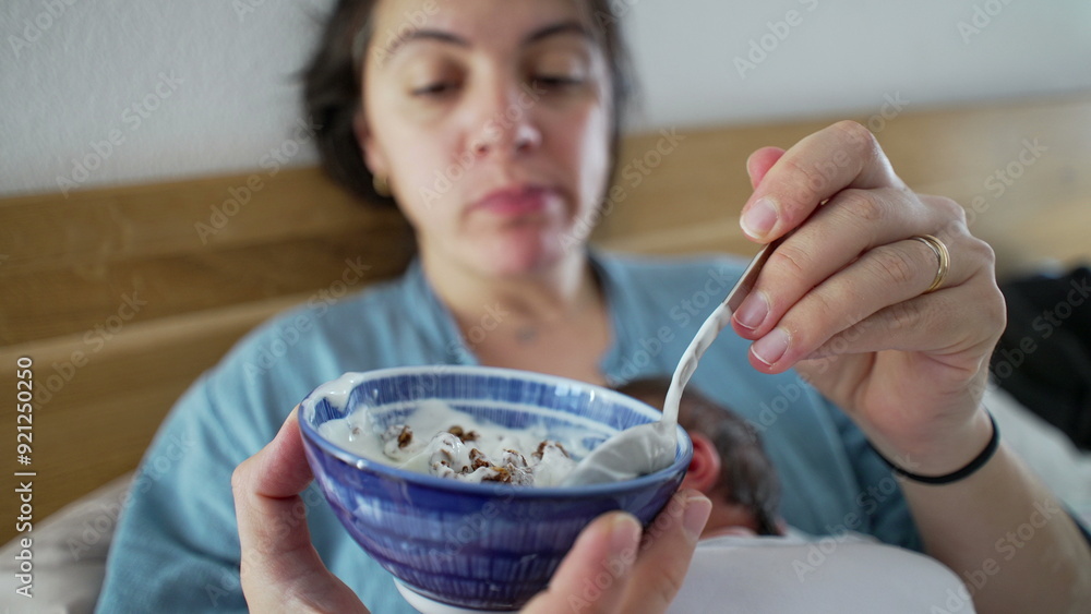 Mother holding bowl of yogurt while newborn baby sleeps on her chest, multitasking parent enjoying snack time, cozy indoor setting, intimate family moment, combining care and nourishment