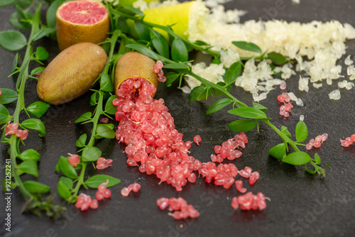 Ripe yellow and burgundy oblong fruits and delicious pink and white citrus caviar on the black slate slab decorated with a green plants sprig. Outlandish Australian finger lime plant indoor growing