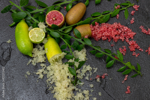 Ripe yellow and burgundy oblong fruits and delicious pink and white citrus caviar on the black slate slab decorated with a green plants sprig. Outlandish Australian finger lime plant indoor growing photo