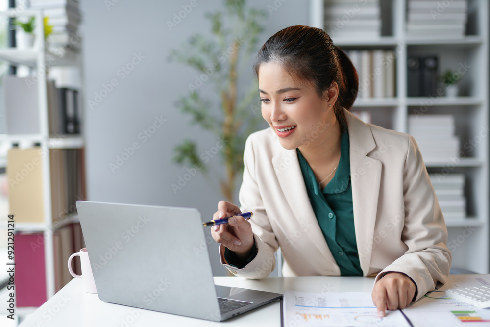 Young asian businesswoman sitting at desk in modern office, analyzing financial charts on laptop and having video call Young asian businesswoman sitting at desk in modern office, analyzing financial charts on laptop and having video call