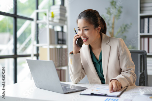 Asian businesswoman is smiling while talking on the phone and analyzing financial charts on a laptop