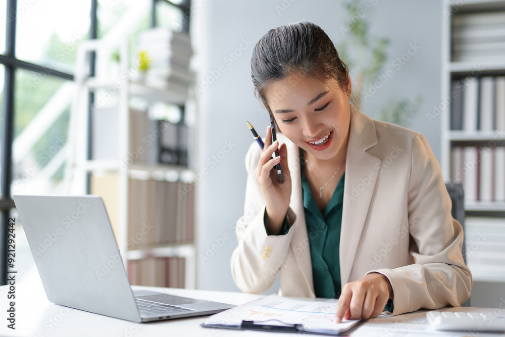 Businesswoman smiling while talking on the phone and pointing at documents during her workday at the office Businesswoman smiling while talking on the phone and pointing at documents during her workday at the office