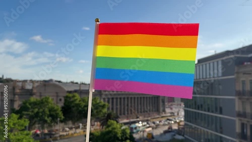 Rainbow flag fluttering in the wind on a balcony with the streets of Barcelona in the background.