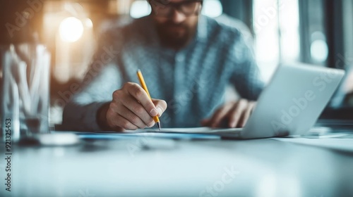 A person sits at a desk using a laptop and takes notes with a pencil on paper, surrounded by office supplies, suggesting productivity and focus in a professional setting. photo