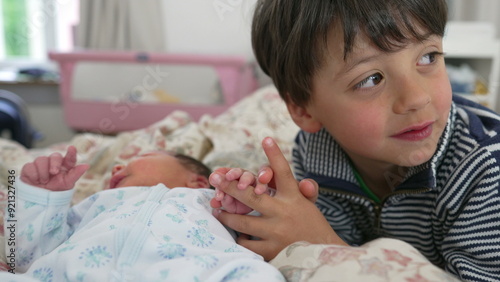 Young boy holding newborn baby’s tiny hand, indoor bedroom setting, gentle interaction, sibling bond, expressions of love and protection, family moment, new beginnings