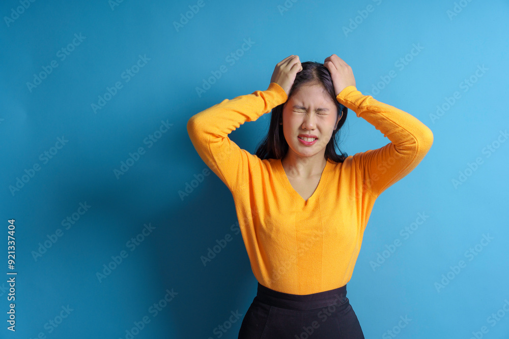Stressed woman pulling hair, feeling frustrated and overwhelmed, with a blue background showing intense emotions. She wears a yellow shirt Stressed woman pulling hair, feeling frustrated and overwhelmed, with a blue background showing intense emotions. She wears a yellow shirt