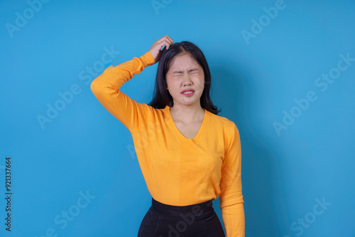 Young woman is scratching her head with a confused expression on her face, standing against a blue background