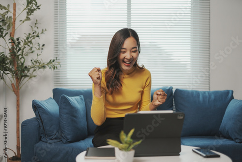 Excited young asian woman sitting on a blue sofa in her living room, raising her fists in the air as she celebrates a success while working from home