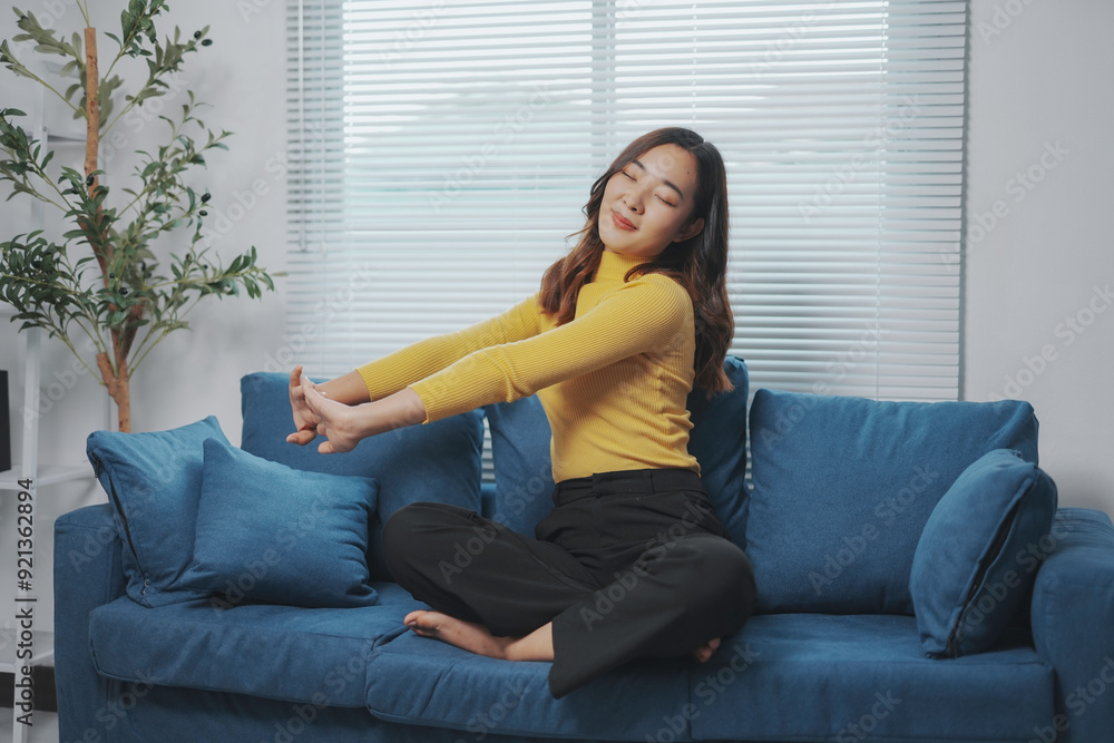 Young woman is sitting cross legged on a sofa in her living room and stretching her arms with her eyes closed Young woman is sitting cross legged on a sofa in her living room and stretching her arms with her eyes closed