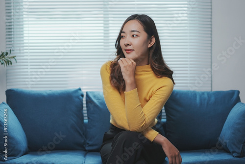 Young asian woman sitting on a sofa is deep in thought, her hand gently resting on her chin as she contemplates something in the distance