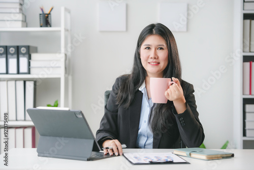 Smiling asian businesswoman drinking coffee while working on a project on a digital tablet in her office