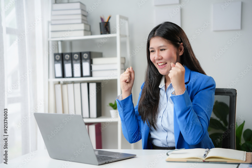 Excited businesswoman raising her fists in front of laptop, celebrating business success in her bright office Excited businesswoman raising her fists in front of laptop, celebrating business success in her bright office