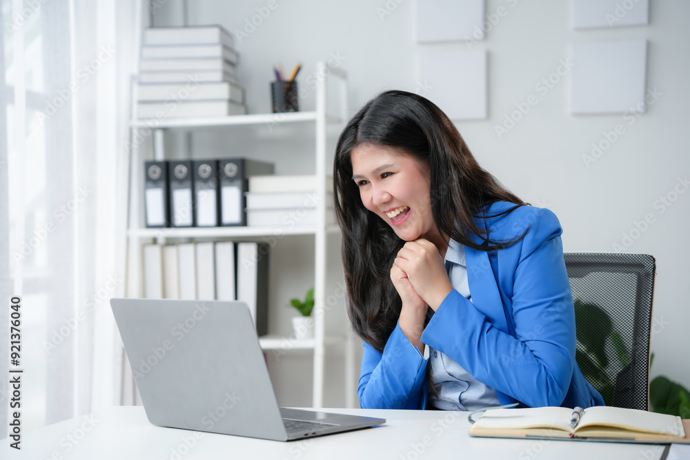 Asian businesswoman is working in her bright office, looking excitedly at her laptop screen and holding her hands together in joy Asian businesswoman is working in her bright office, looking excitedly at her laptop screen and holding her hands together in joy