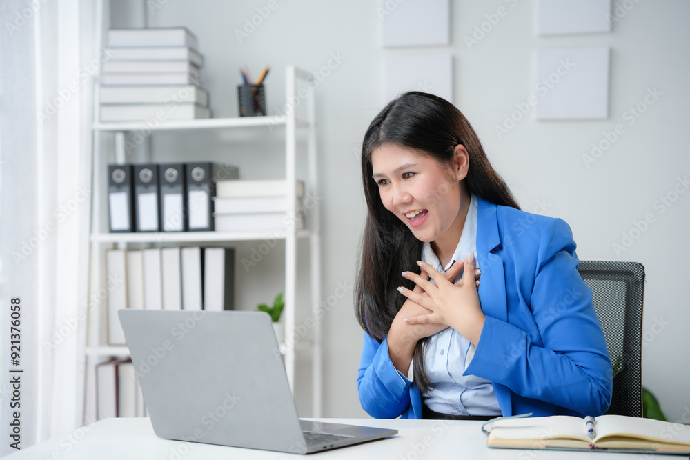Asian businesswoman is holding her chest and feeling touched while reading an email on a laptop in her bright office Asian businesswoman is holding her chest and feeling touched while reading an email on a laptop in her bright office