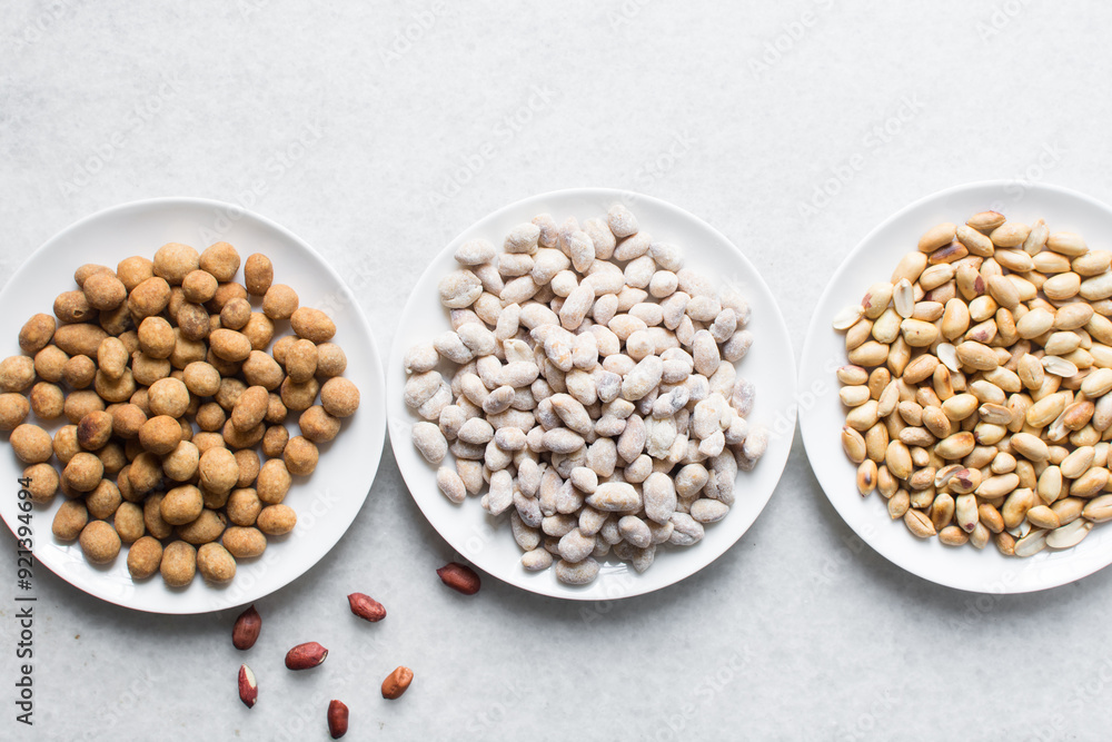 Overhead view of roasted peanuts and coated peanuts on a white plate, flatlay of roasted peeled peanuts and raw coated peanuts on a marble countertop, process of making coated nuts or cracker nuts Overhead view of roasted peanuts and coated peanuts on a white plate, flatlay of roasted peeled peanuts and raw coated peanuts on a marble countertop, process of making coated nuts or cracker nuts