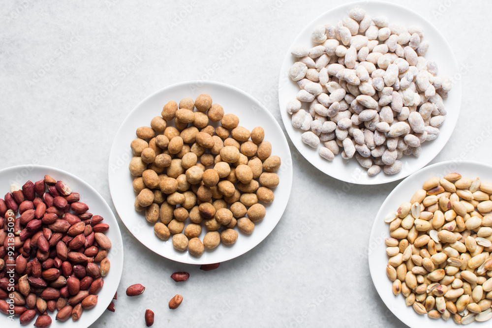 Overhead view of roasted peanuts and coated peanuts on a white plate, flatlay of roasted peeled peanuts and raw coated peanuts on a marble countertop, process of making coated nuts or cracker nuts Overhead view of roasted peanuts and coated peanuts on a white plate, flatlay of roasted peeled peanuts and raw coated peanuts on a marble countertop, process of making coated nuts or cracker nuts