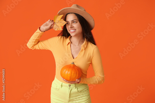 Stylish young woman with fresh pumpkin and autumn leaf on orange background