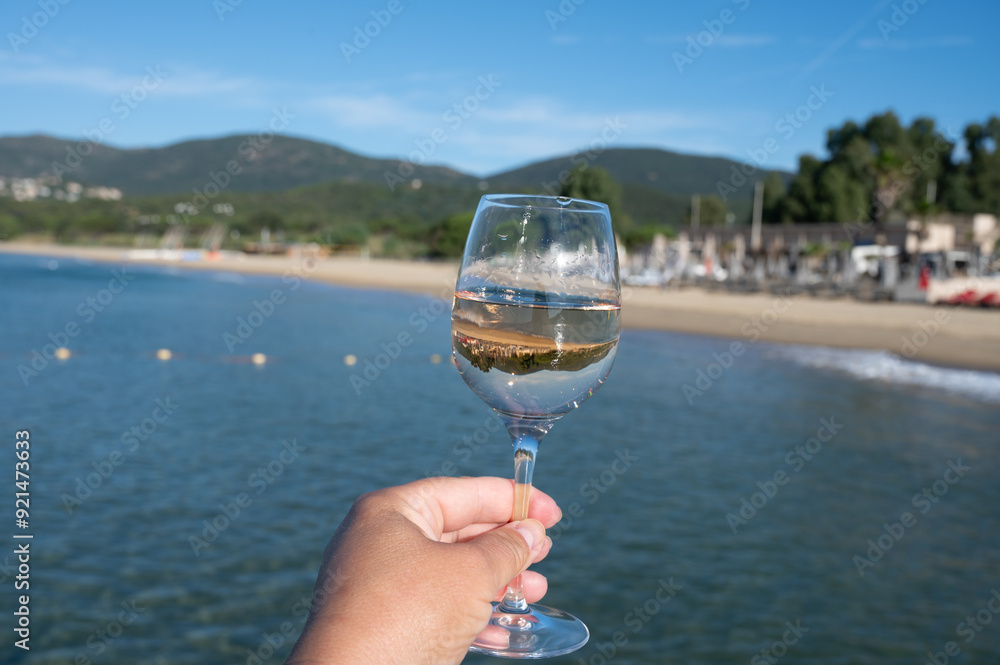 Obraz premium Hand with glass of cold rose wine from Provence and wooden yacht boota pier on white sandy beach Plage de Pampelonne near Saint-Tropez, summer vacation in France