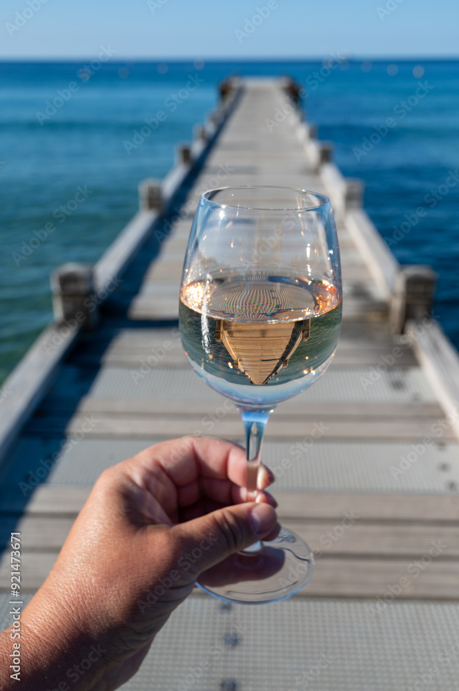 Obraz premium Hand with glass of cold rose wine from Provence and wooden yacht boota pier on white sandy beach Plage de Pampelonne near Saint-Tropez, summer vacation in France