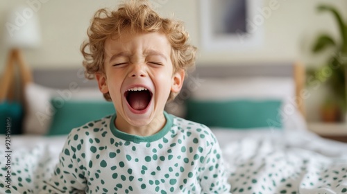 A young boy with curly hair passionately shouting while wearing green polka dot pajamas, sitting on a bed in a bright and cozy bedroom, expressing excitement and energy. photo