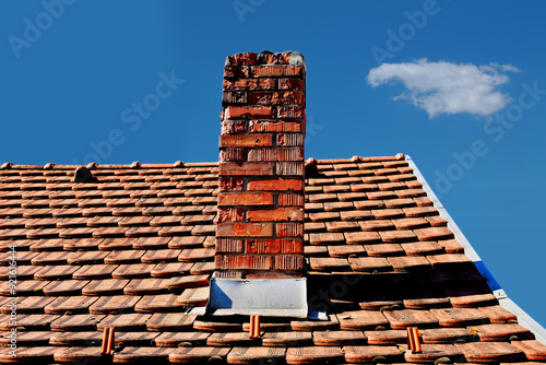 damaged clay brick chimney with weathered and spalling surface. clay tile roofing. gray metal flashing. strong shadow. blue sky background. summer scene. building, home maintenance and upkeep concept