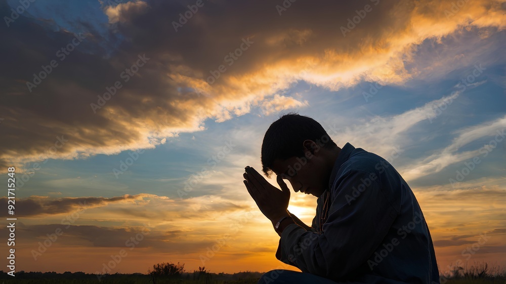 Fototapeta premium A young man’s silhouette on his knees, praying with clasped hands, set against a vibrant sunset, symbolizing sacrifice and faith.