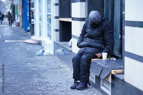 Anonymous homeless man wearing black clothes on a cold winter day standing on a windowsill on cardboard boxes with a white cup of hot drink coffee or tea in front of him