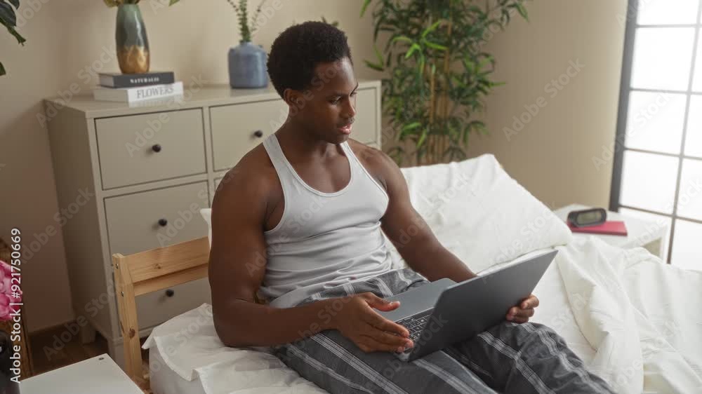Young man in a bedroom using a laptop for a video call while sitting on a bed next to a dresser and plants, wearing a white tank top and striped pants Young man in a bedroom using a laptop for a video call while sitting on a bed next to a dresser and plants, wearing a white tank top and striped pants