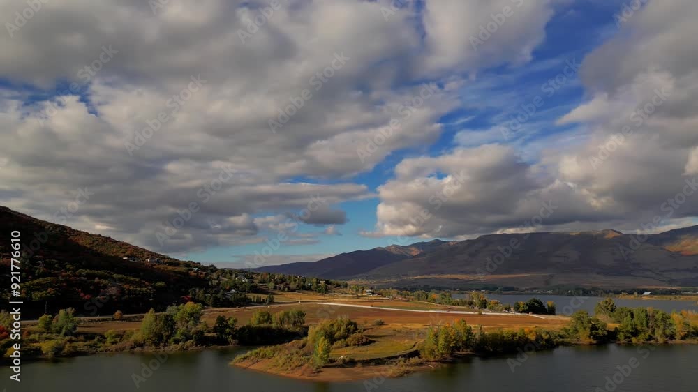 Moving drone shot of a beautiful lake reservoir and vast tree plagued mountains in the Fall, with orange, red and yellow trees and bright, cloudy blue sky. Moving drone shot of a beautiful lake reservoir and vast tree plagued mountains in the Fall, with orange, red and yellow trees and bright, cloudy blue sky.