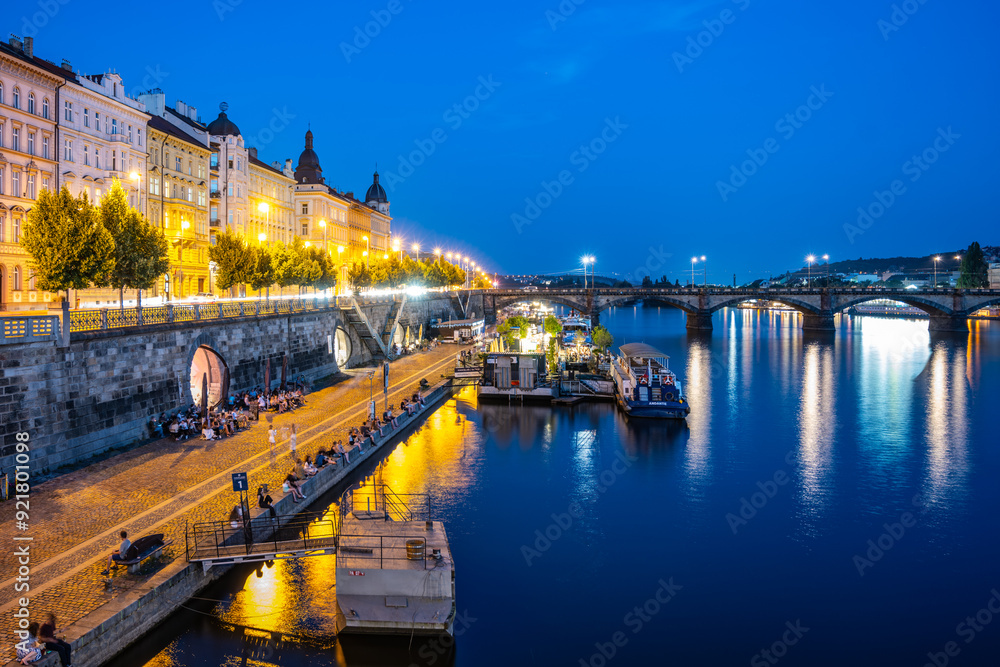 Fototapeta premium The Vltava River in Prague is bustling with activity at night. People are gathered on the embankment, enjoying the cool evening air. The city lights reflect off the water, creating a beautiful and