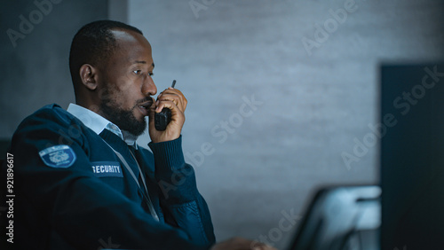 Canvas Print African American security officer or dispatch in uniform uses tablet computer and walkie talkie, looks at screens working in monitoring center during night shift