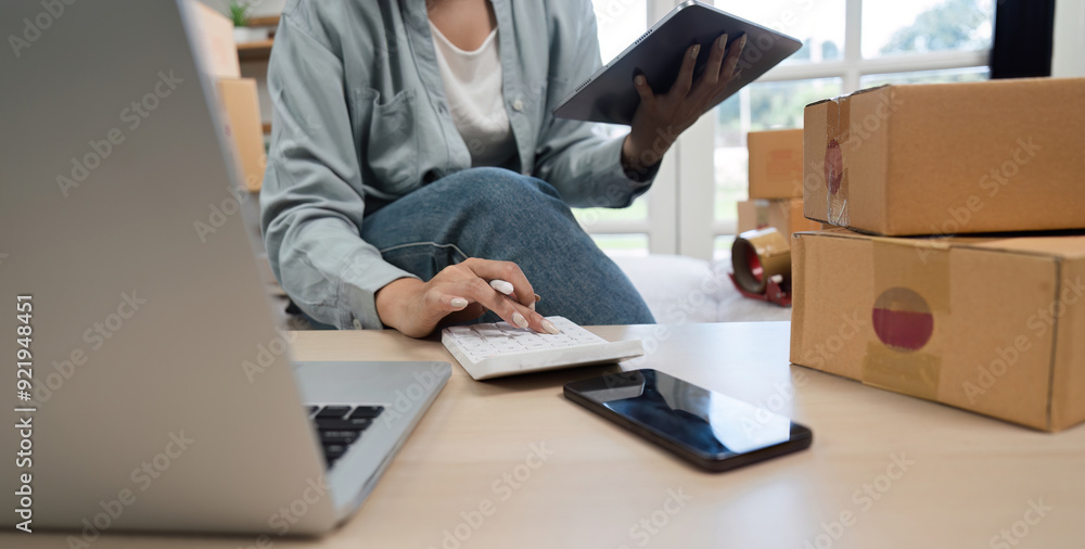 Young Woman Managing Online Business from Home, Using Tablet and Calculator, Surrounded by Shipping Boxes and Laptop Young Woman Managing Online Business from Home, Using Tablet and Calculator, Surrounded by Shipping Boxes and Laptop